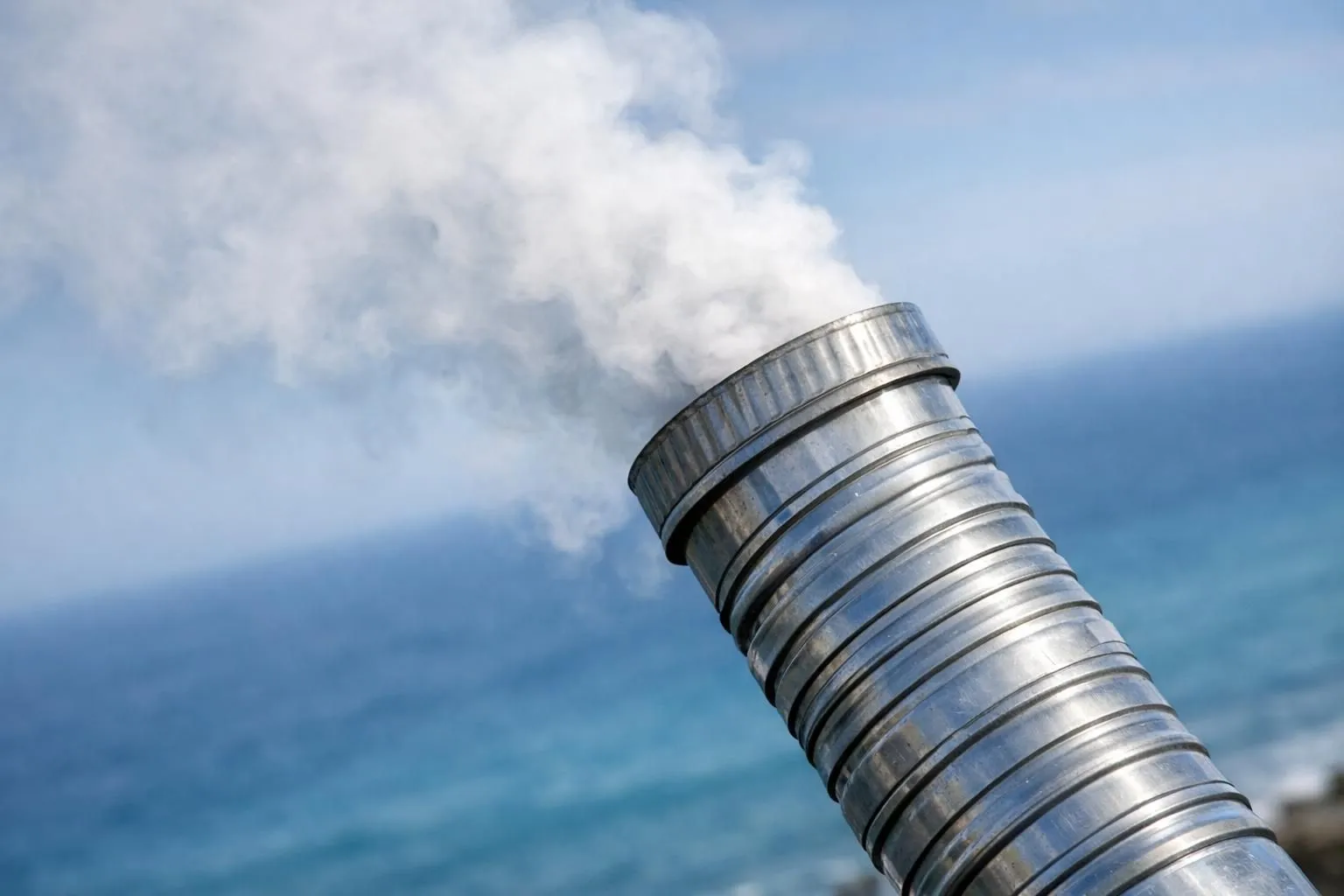 Metallic exhaust pipe emitting smoke against cloudy blue sky.